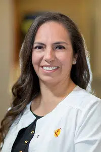 The image features a smiling woman with dark hair wearing a white lab coat, standing against a blurred background.