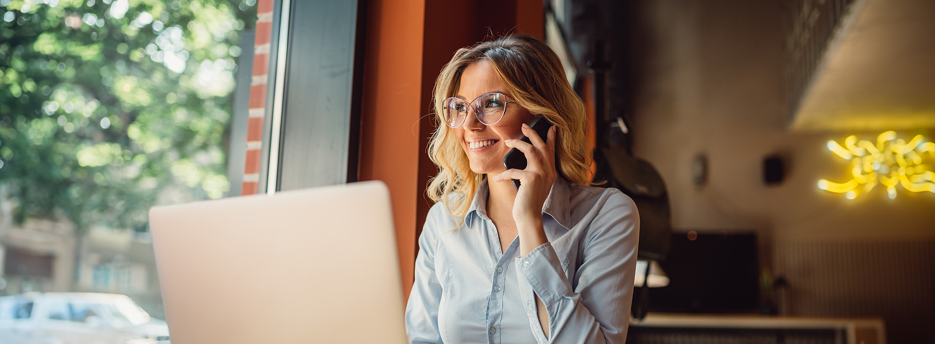A woman sitting at a laptop in a cafe setting, engaged in a phone call while working.