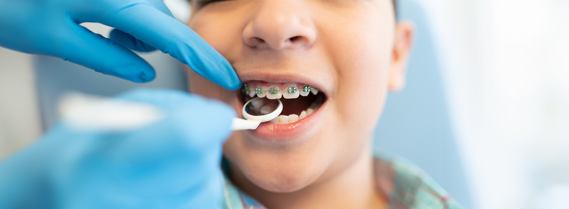 The image shows a young boy receiving dental care with a dental hygienist, wearing gloves and using a dental mirror to examine his teeth.