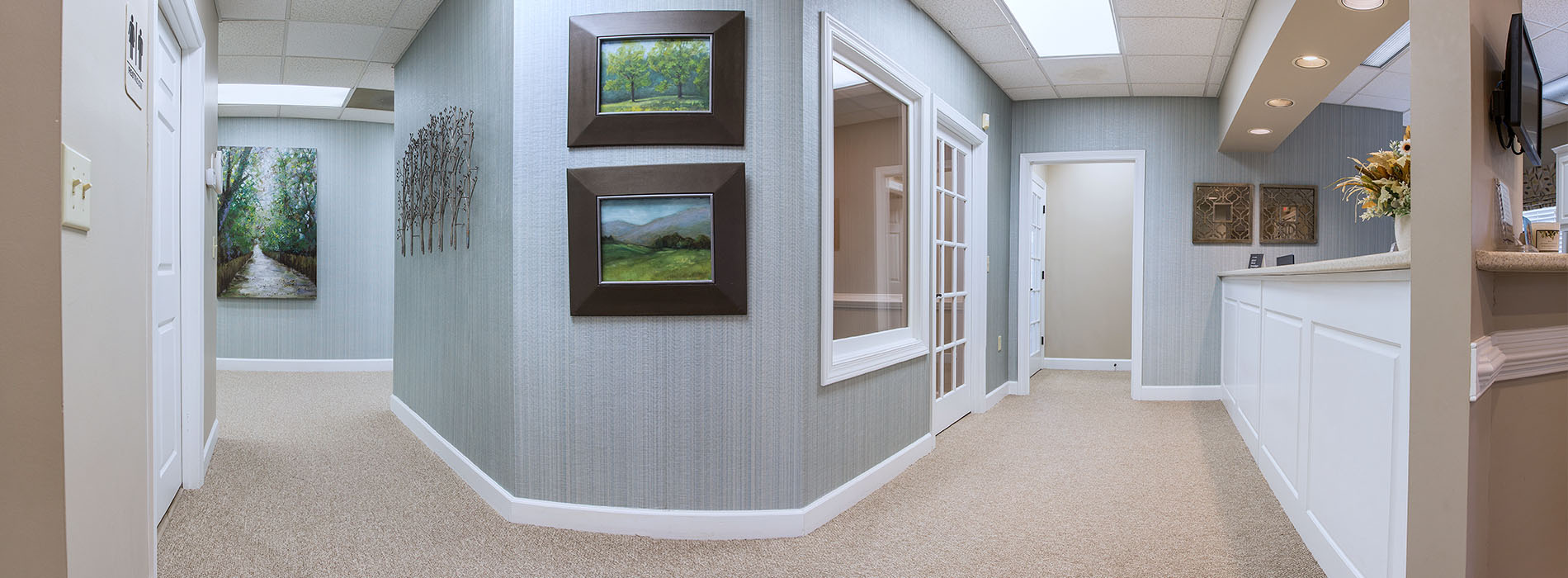The image shows an interior space that appears to be a waiting room or hallway within a medical office, featuring a wall with artwork, a white door on the right, and a counter with a framed picture on top.