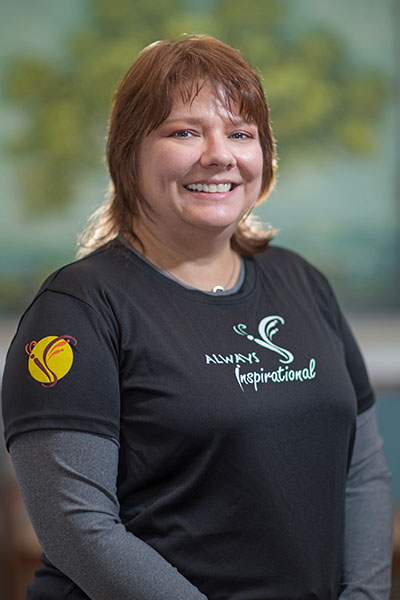 The image shows a person smiling at the camera, wearing a black shirt with a logo on the front, and posing against a background with a framed picture.